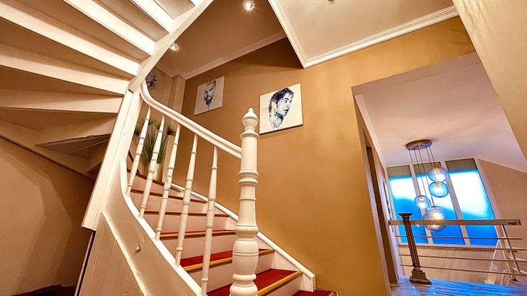 Un escalier élégant avec une balustrade blanche et des tapis rouges monte. Des images sont accrochées aux murs, et à l'arrière-plan, de grandes fenêtres sont visibles.