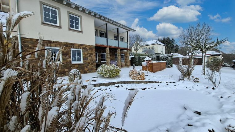 Une jolie maison avec un jardin recouvert de neige. Le ciel est dégagé et il y a quelques nuages.