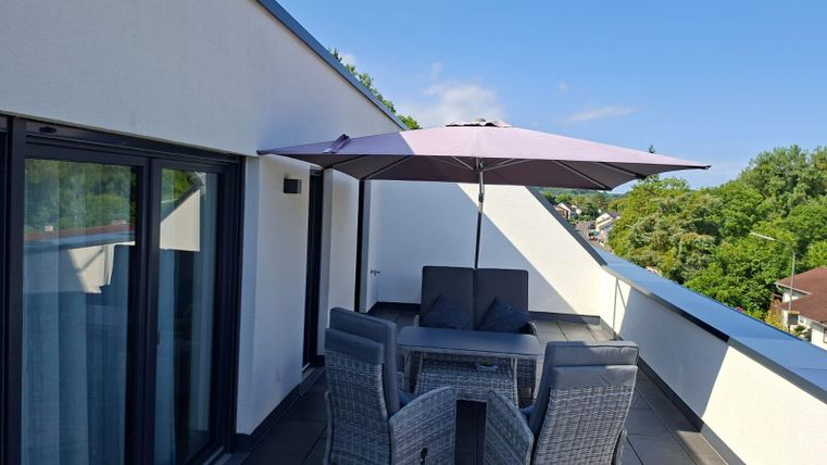 A modern rooftop terrace with a dining table and chairs. A sun umbrella provides shade, surrounded by green trees and a clear sky.
