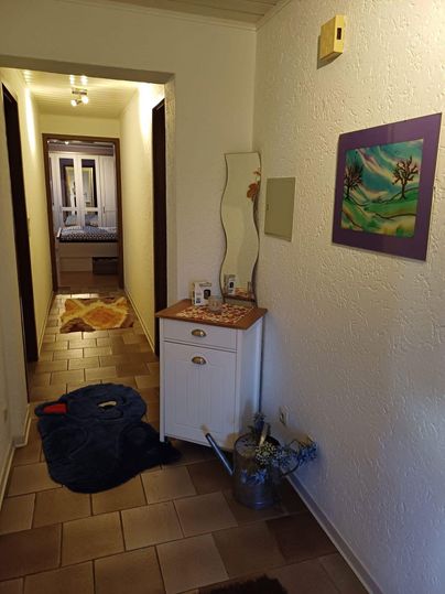 Ein schmaler Flur mit einem kleinen Tisch und einem Spiegel. An der Wand hängt ein Gemälde, der Boden ist mit Fliesen ausgelegt.