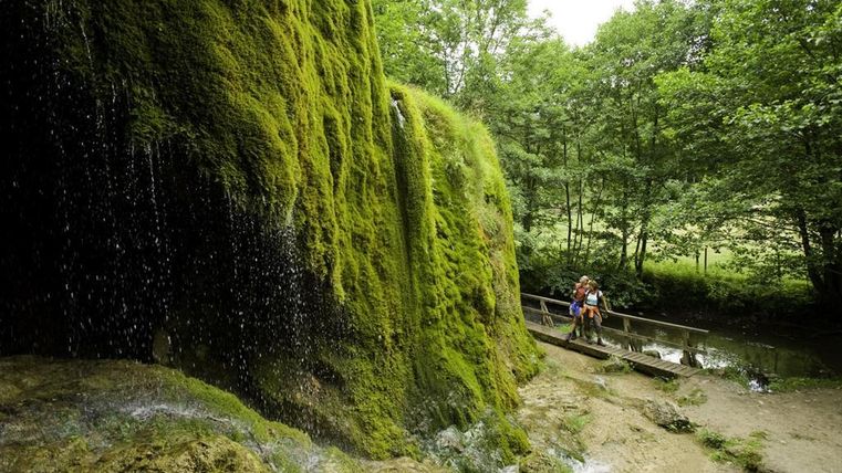 Een groene, met mos bedekte rotswand met een kleine waterval. Op de voorgrond zijn er mensen op een houten brug die de rustige rivier oversteken.
