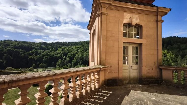 Een historisch gebouw met een open terras en grote ramen. Op de achtergrond strekt een bos zich uit onder een heldere blauwe lucht.