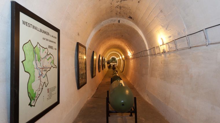 Ein langer, heller Tunnel mit gezeichneten Karten an den Wänden. In der Mitte stehen historische militärische Objekte, die den kühlen Charakter des Raumes unterstreichen.