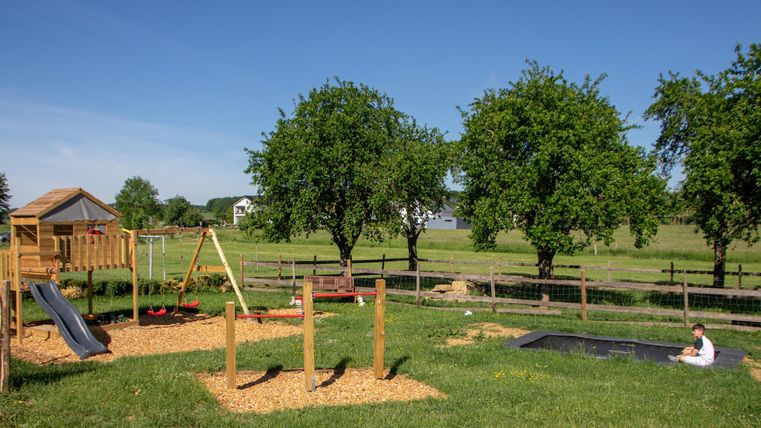 Ein Spielplatz mit Rutsche, Schaukeln und einem Trampolin in einer grünen Wiese. Im Hintergrund gibt es einige Bäume und ein ländliches Landschaftsbild.
