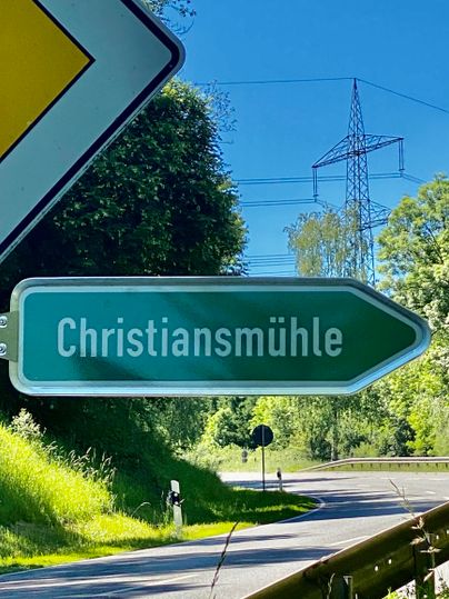 Ein Verkehrsschild zeigt die Richtung nach Christiansmühle. Im Hintergrund sind Bäume und ein blauer Himmel sichtbar.
