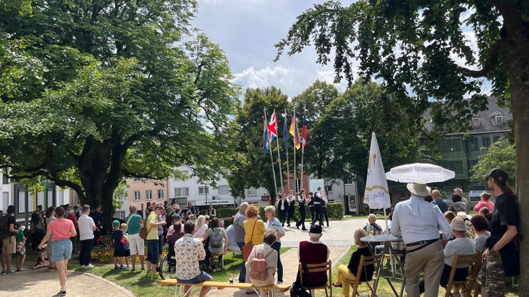Eine Gruppe von Menschen versammelt sich in einem Park, um eine Aufführung zu sehen. Bunte Flaggen sind aufgestellt, und es herrscht eine festliche Atmosphäre.