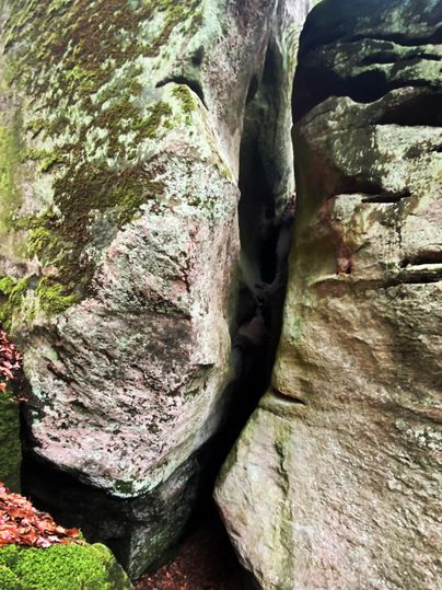 Ein schmaler Spalt zwischen großen, moosbedeckten Felsen im Wald. Die Umgebung ist grün und wirkt natürlich.