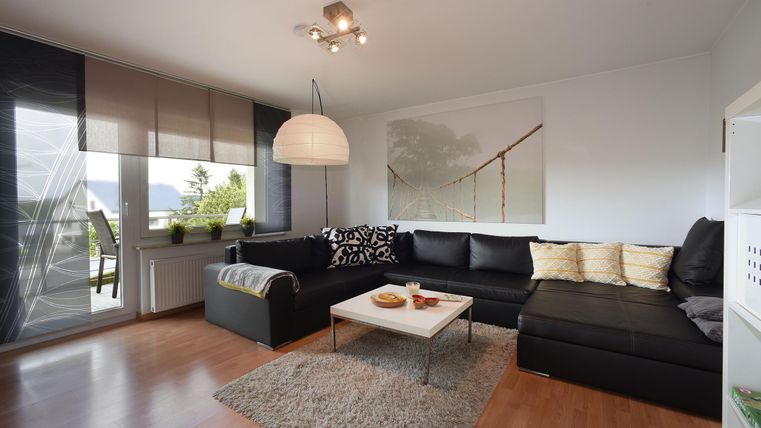 Un salon moderne avec un canapé noir, un tapis clair et une table garnie de collations. De grandes fenêtres laissent entrer beaucoup de lumière naturelle.