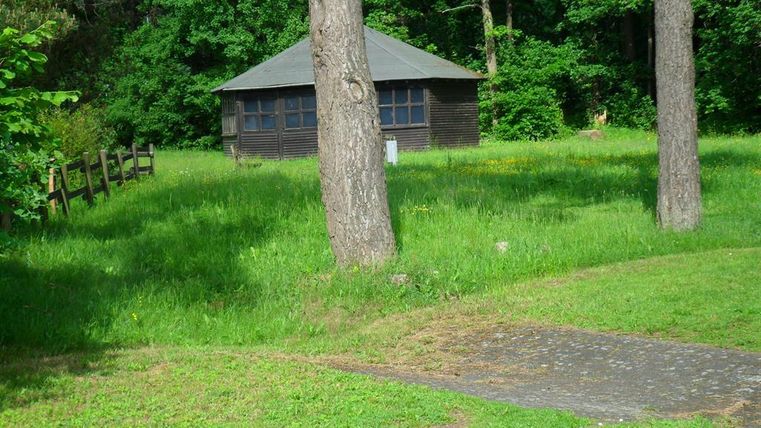 Une simple maison en bois se dresse entourée de verdure luxuriante et d'arbres. Le sol est recouvert d'herbe et d'un petit chemin pavé.