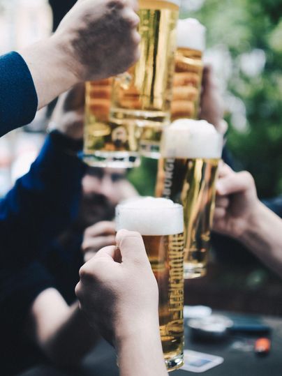 Angehobene Biergläser werden von mehreren Personen gestoßen. Der Hintergrund ist verschwommen und es herrscht eine gesellige Stimmung.