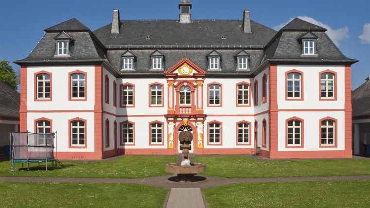 Ein historisches Gebäude mit roter und weißer Fassade, umgeben von Rasen. Der klare blaue Himmel und die Symmetrie des Gebäudes verleihen ihm ein imposantes Aussehen.