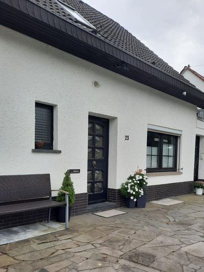 A modern house with a white facade and a dark roof. In front of the door, there is a bench and blooming plants.