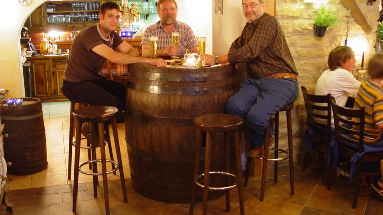 Trois hommes sont assis à une table ronde en fût de bois dans un bar accueillant. En arrière-plan, des étagères avec des boissons et une atmosphère conviviale sont visibles.