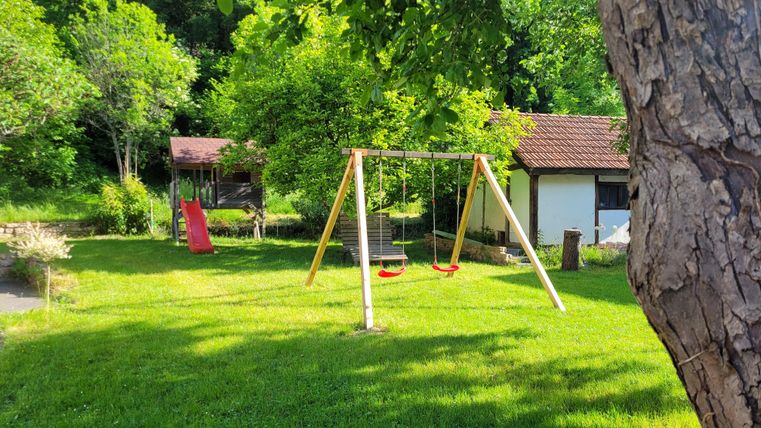 Ein Spielplatz mit einer Schaukel und einer Rutsche in einem grünen Garten. Im Hintergrund ist ein kleines Haus und viele Bäume zu sehen.