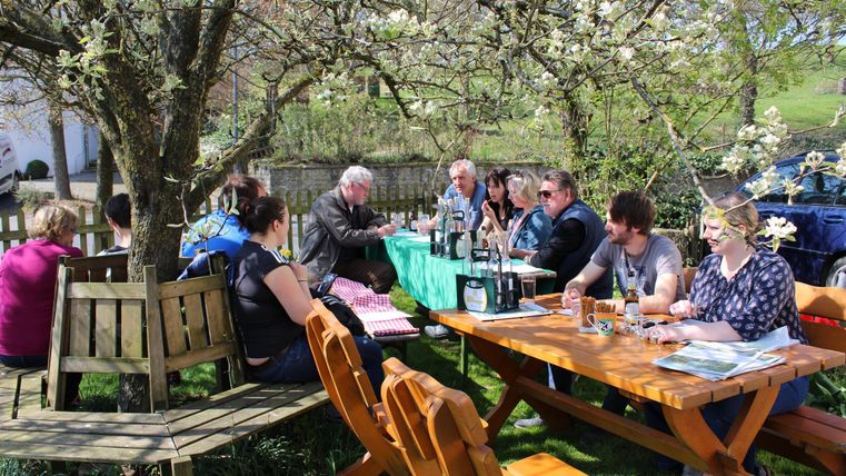 Een groep mensen zit aan tafels in de buitenlucht onder een bloeiende boom. Het lijkt een zonnige dag te zijn, ideaal voor een ontmoeting in de buitenlucht.