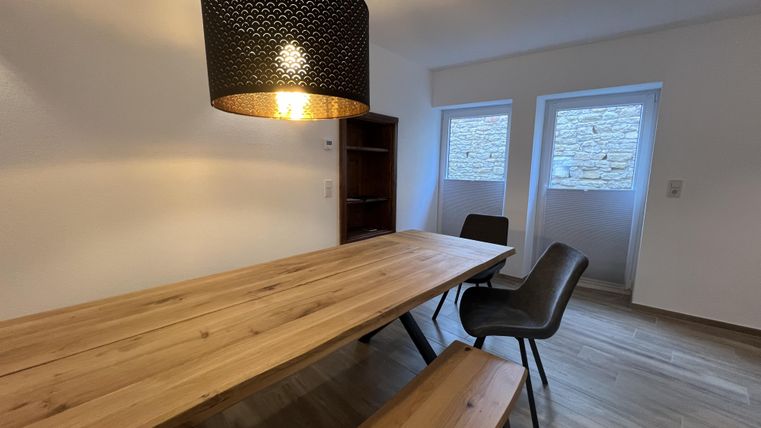 Een lichte eetruimte met een lange houten tafel en moderne stoelen. Boven de tafel hangt een opvallende, zwarte lamp.