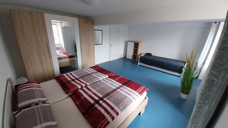 Ein modernes Schlafzimmer mit einem großen Doppelbett und einem Einzelbett. Der Raum ist hell und freundlich, mit einem blauen Fußboden und minimaler Einrichtung.