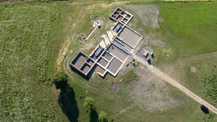 Eine Baustelle mit mehreren Gebäudestrukturen und Wegen. Das Gelände ist umgeben von Wiesen und Feldern.