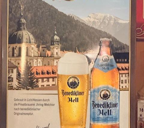 Een reclamebord voor Benediktiner Hell Bier. De afbeelding toont een biertje en een fles voor een schilderachtig berglandschap.