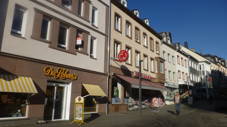 Eine ruhige Straßenszene mit mehreren Geschäften und Häusern. Die Sonne scheint und es gibt wenig Verkehr.