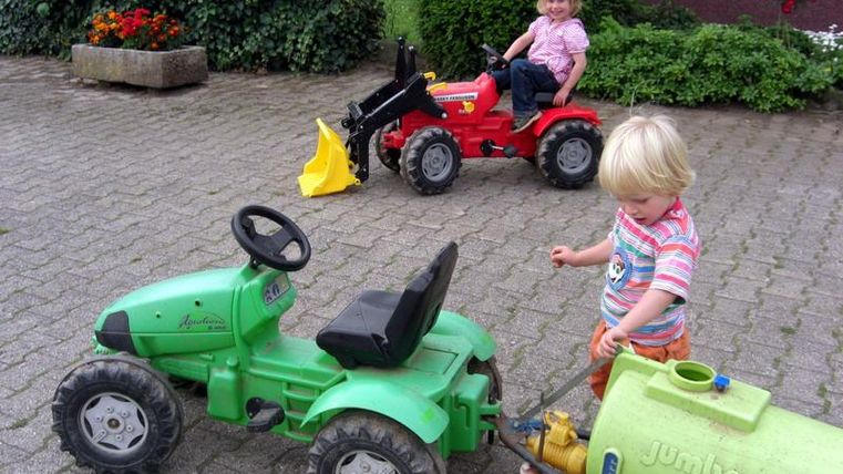 Deux enfants jouent avec des mini-tracteurs sur une cour pavée. L'un des enfants conduit un tracteur vert, tandis que l'autre est assis sur un tracteur rouge.