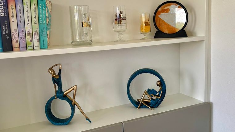 Eine moderne Regal mit dekorativen Skulpturen und Gläsern. Im Hintergrund stehen Bücher und eine Uhr, die eine stilvolle Atmosphäre schaffen.