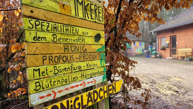 Een kleurrijk houten bord met informatie over bijenproducten staat voor een klein verkoopwinkeltje. De omgeving is gekenmerkt door bomen en herfstbladeren.