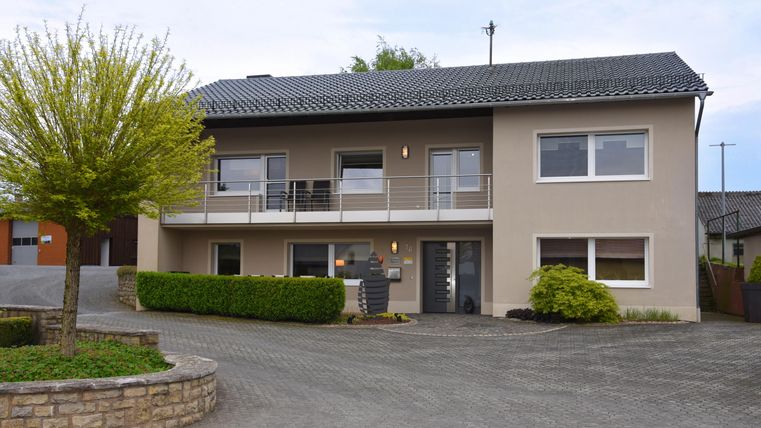 Een modern huis met een balkonreling en een verzorgde voortuin. De gevel is in zachte kleuren gehouden en het terrein is goed aangelegd.