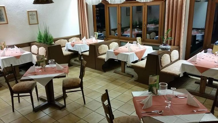Ein gemütliches Restaurant mit Tischen und Stühlen. Die Tische sind mit weißen und roten Tischdecken gedeckt und es gibt Pflanzen im Hintergrund.