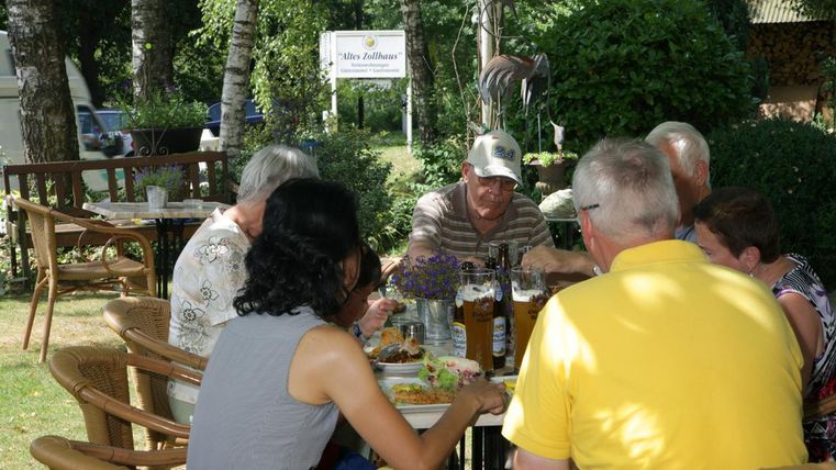 Un groupe de personnes est assis à l'extérieur et profite d'un repas ensemble. Entouré d'arbres et d'une ambiance accueillante, la scène semble amicale et conviviale.