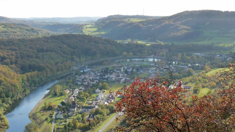 A picturesque landscape with green hills and a river winding through the valley. In the foreground, there is a small village with residential houses and colorful trees.