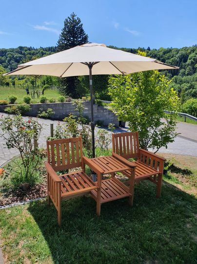 Twee houten stoelen onder een parasol staan in een groene tuin. Op de achtergrond zijn bomen en struiken te zien.