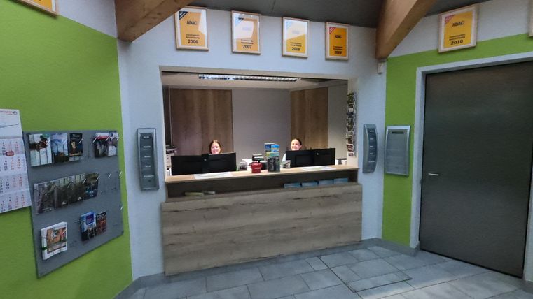 Un espace d'accueil avec deux employées souriantes derrière un comptoir d'accueil moderne. Les murs sont peints dans des couleurs fraîches, et il y a des documents d'information sur le côté.