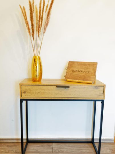 Une table en bois moderne avec un cadre noir. Sur la table se trouve un vase jaune avec des fleurs séchées et un livre d'invités.