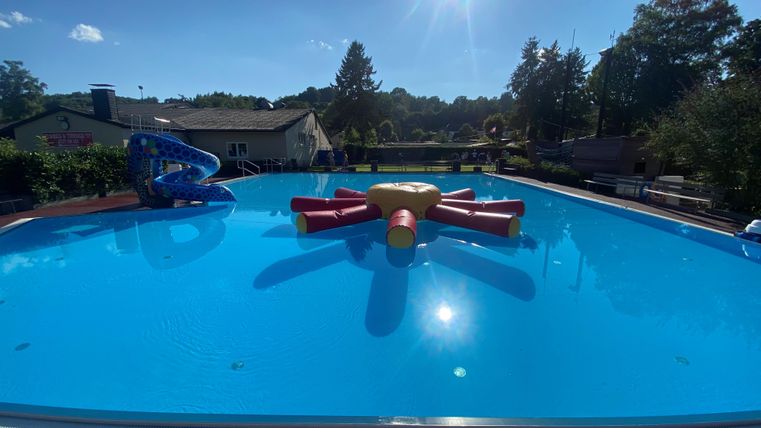 Een groot zwembad met helderblauw water en een kleurrijk, opblaasbaar speelgoed in het midden. Op de achtergrond zijn bomen en een gebouw te zien.