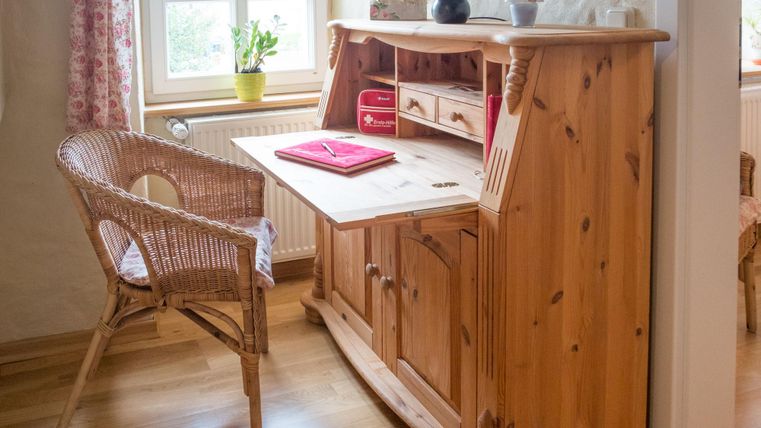 Een houten bureau met een rieten stoel staat in een lichte kamer. Op de tafel ligt een rood notitieboek en voor het raam staat een plant.
