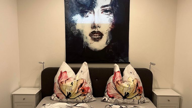 Une chambre moderne avec un lit et des housses de coussin colorées. Un portrait artistique d'une femme est accroché au mur.