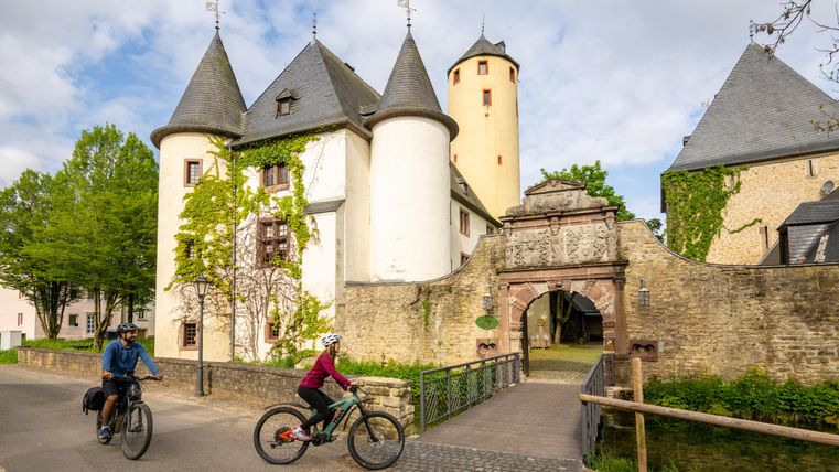 Un château pittoresque avec des tours et une végétation verte. Des cyclistes passent tranquillement sur le chemin menant au château.