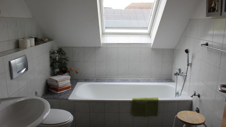 Een moderne badkamer met een bad, een wastafel en een raam. De muren zijn betegeld en er zijn enkele handdoeken en een plant zichtbaar.