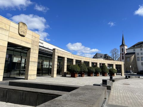 Ein modernes Gebäude mit großen Fenstern und einem klaren Himmel. Im Vordergrund sind Pflanzkübel und ein Brunnen zu sehen.
