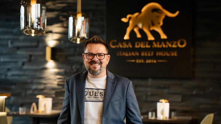 Een glimlachende man staat in een elegant restaurant met de inscriptie "Casa del Manzo" op de achtergrond. De sfeer is modern en uitnodigend.