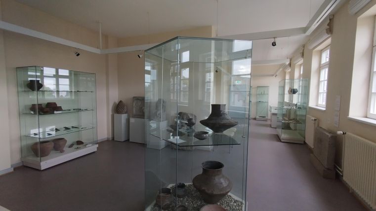A museum with display cases showcasing various clay vessels and archaeological artifacts. The room is bright and has large windows.