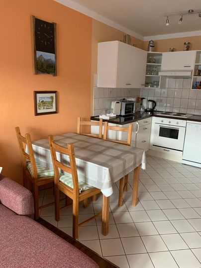 Een lichte keuken met een eetgedeelte en houten stoelen. De muren zijn oranje en er zijn verschillende keukenapparaten.