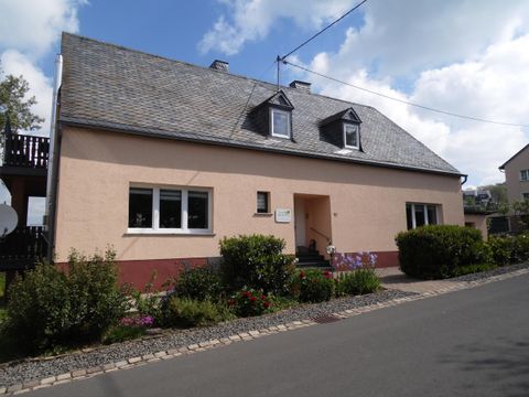 Ein zweigeschossiges, mauerfarbenes Haus mit einem grauen Dach. Es hat große Fenster und ist von gepflegten Pflanzen umgeben.