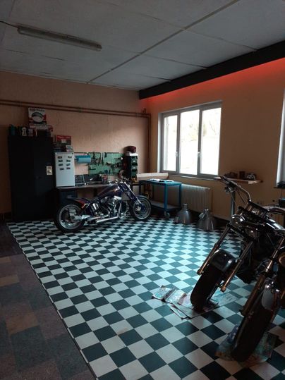 Eine Garage mit einem schachbrettartigen Boden und mehreren Motorrädern. Es gibt ein Fenster, das Tageslicht hereinlässt, und Werkzeuge auf einer Werkbank.