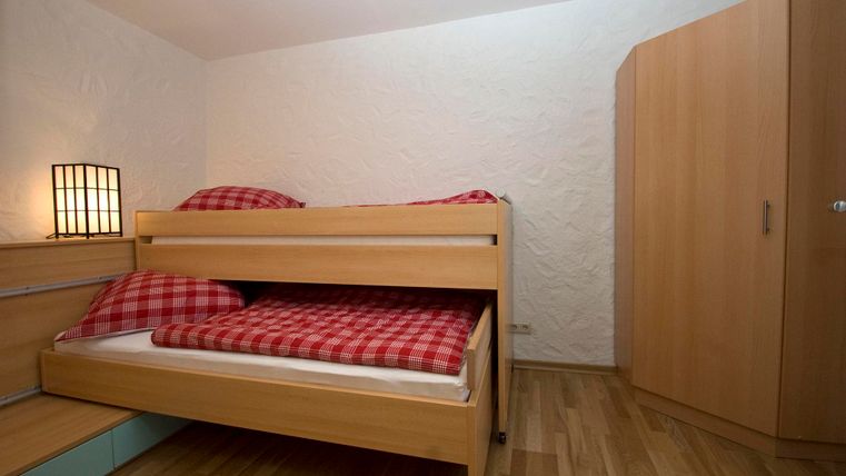 Een eenvoudige slaapkamer met een stapelbed en rode geruite kussens. De ruimte heeft lichte muren en een houten vloer.