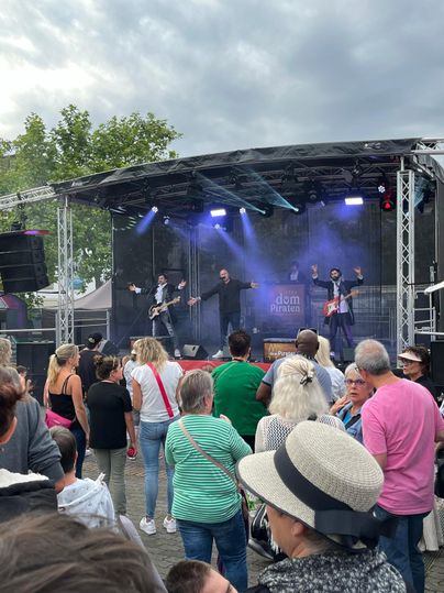 Eine Band spielt auf einer Bühne vor einem begeisterten Publikum. Viele Menschen stehen zusammen und genießen die Musik unter einem bewölkten Himmel.