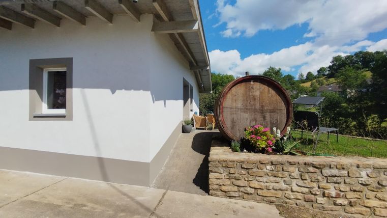 Een modern huis met een lichte gevel en een grote houten schuur ernaast. Op de achtergrond zijn groene heuvels en een blauwe lucht te zien.