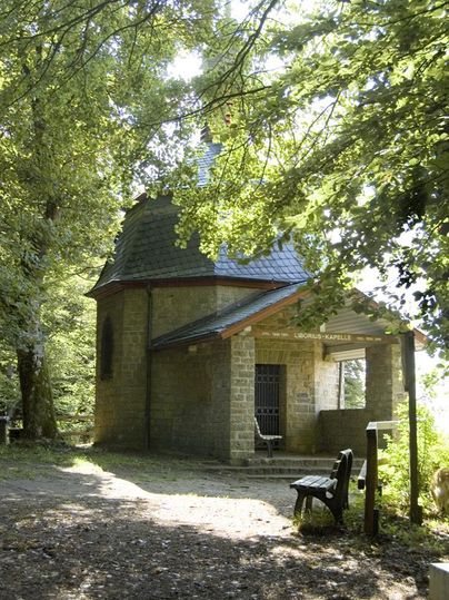 Une petite chapelle entourée d'arbres dans un environnement paisible. Un banc en bois invite à la détente.