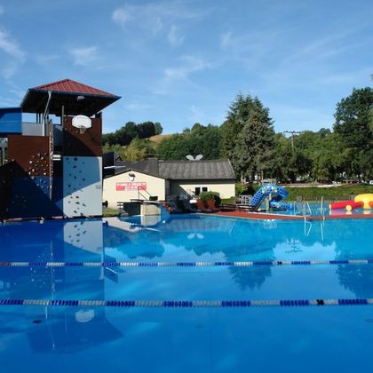 Une piscine avec une surface d'eau bleue et un toboggan. À l'arrière-plan, on aperçoit des espaces verts et des équipements de jeu.